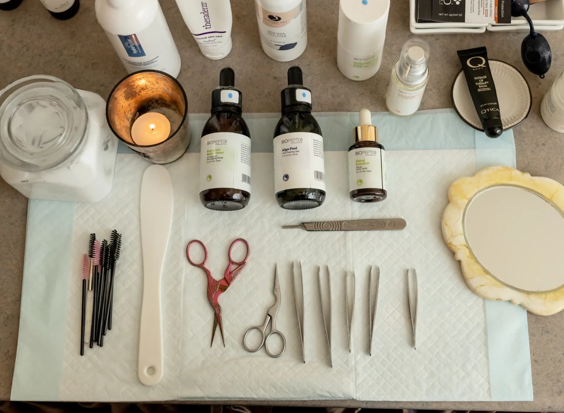 Overhead flatlay of ESSpa facial essentials — amber serum bottles, a lit candle, scissors, tweezers, brushes and a scalloped hand mirror laid out on a fresh treatment towel