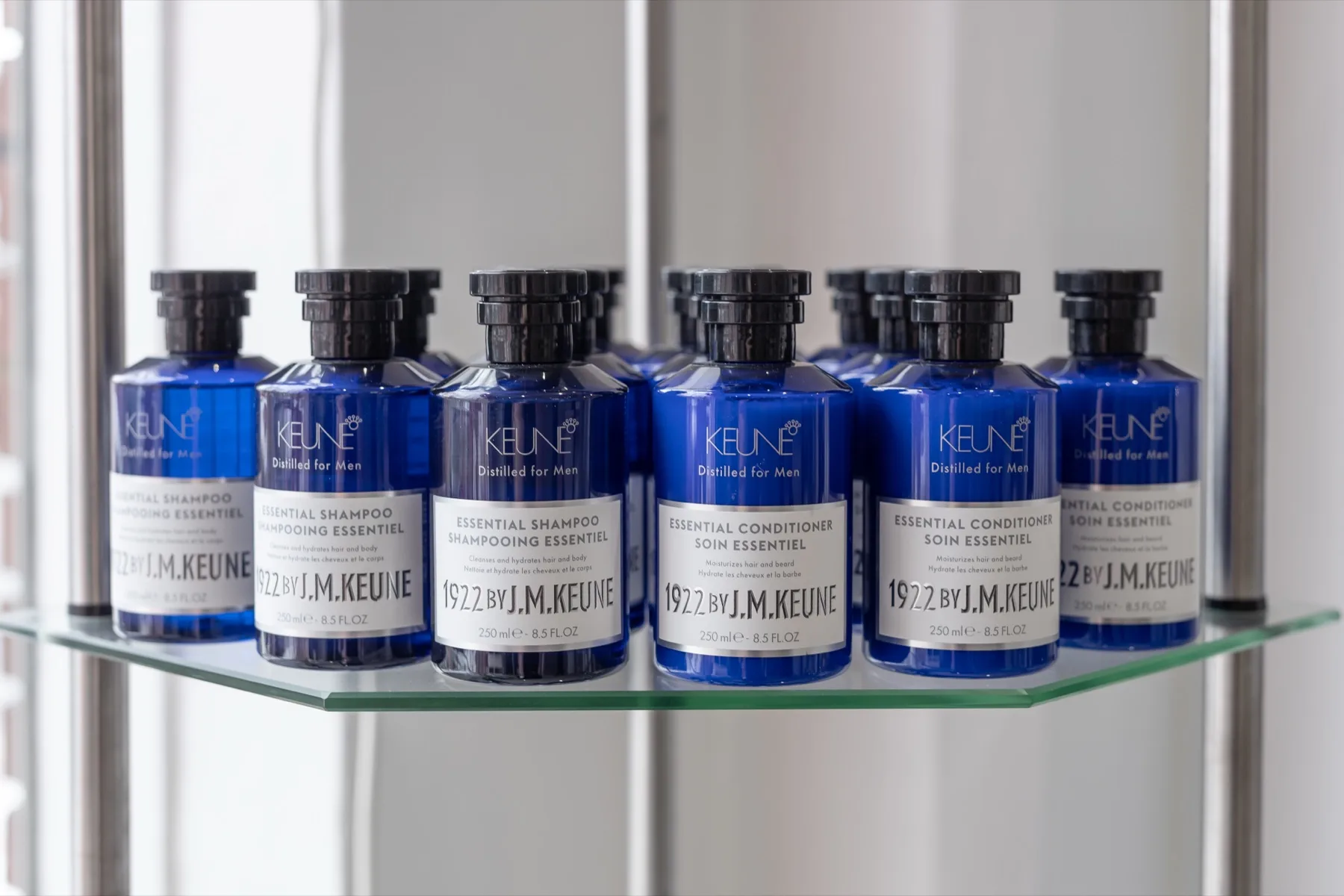 Deep cobalt blue Keune 1922 shampoo and conditioner bottles lined up on a glass shelf in cool, water-clear light.