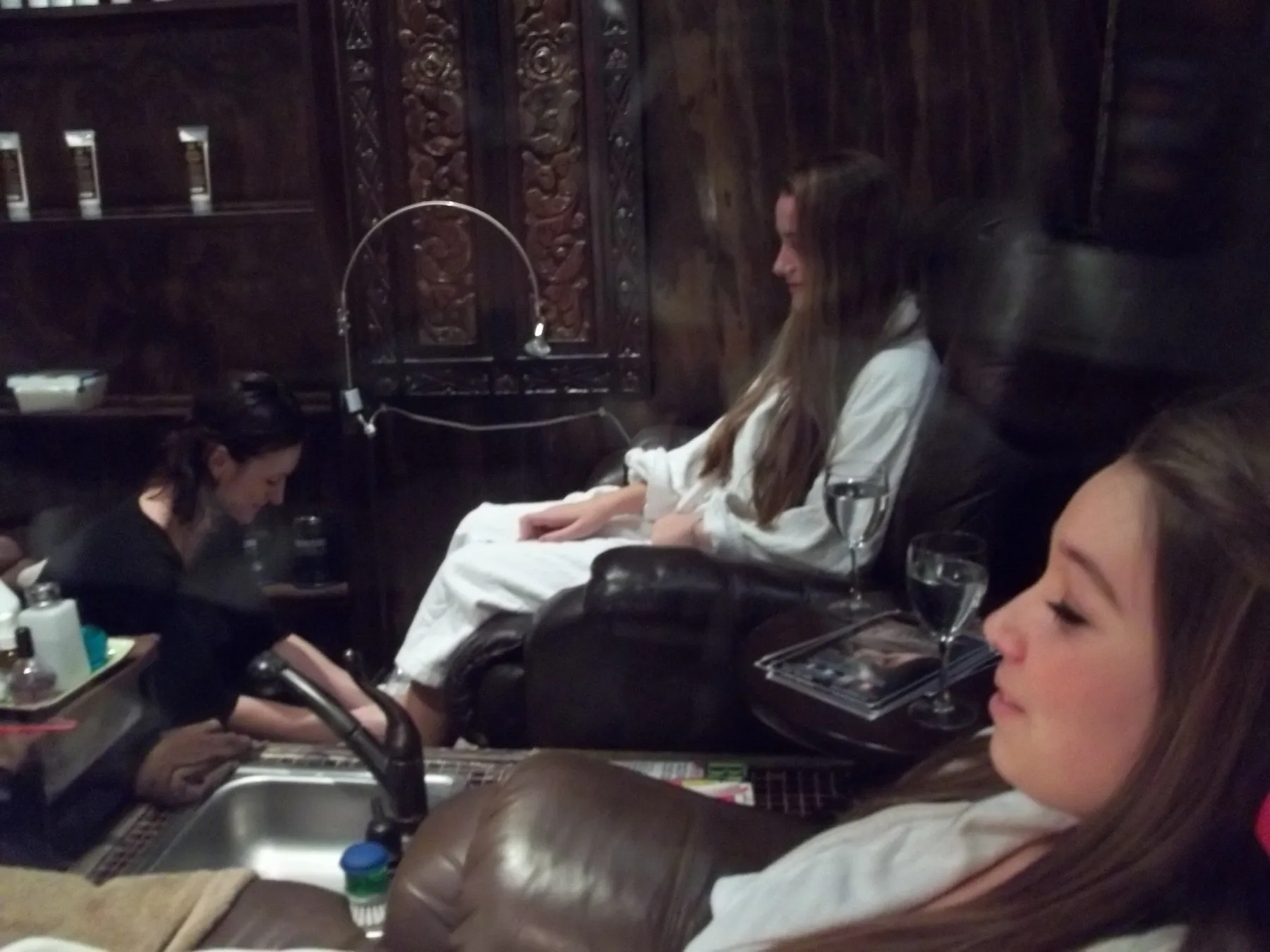 Guests unwinding in white robes in ESSpa's carved-wood pedicure lounge, a technician tending a foot soak at the basin while wine glasses wait on the side table