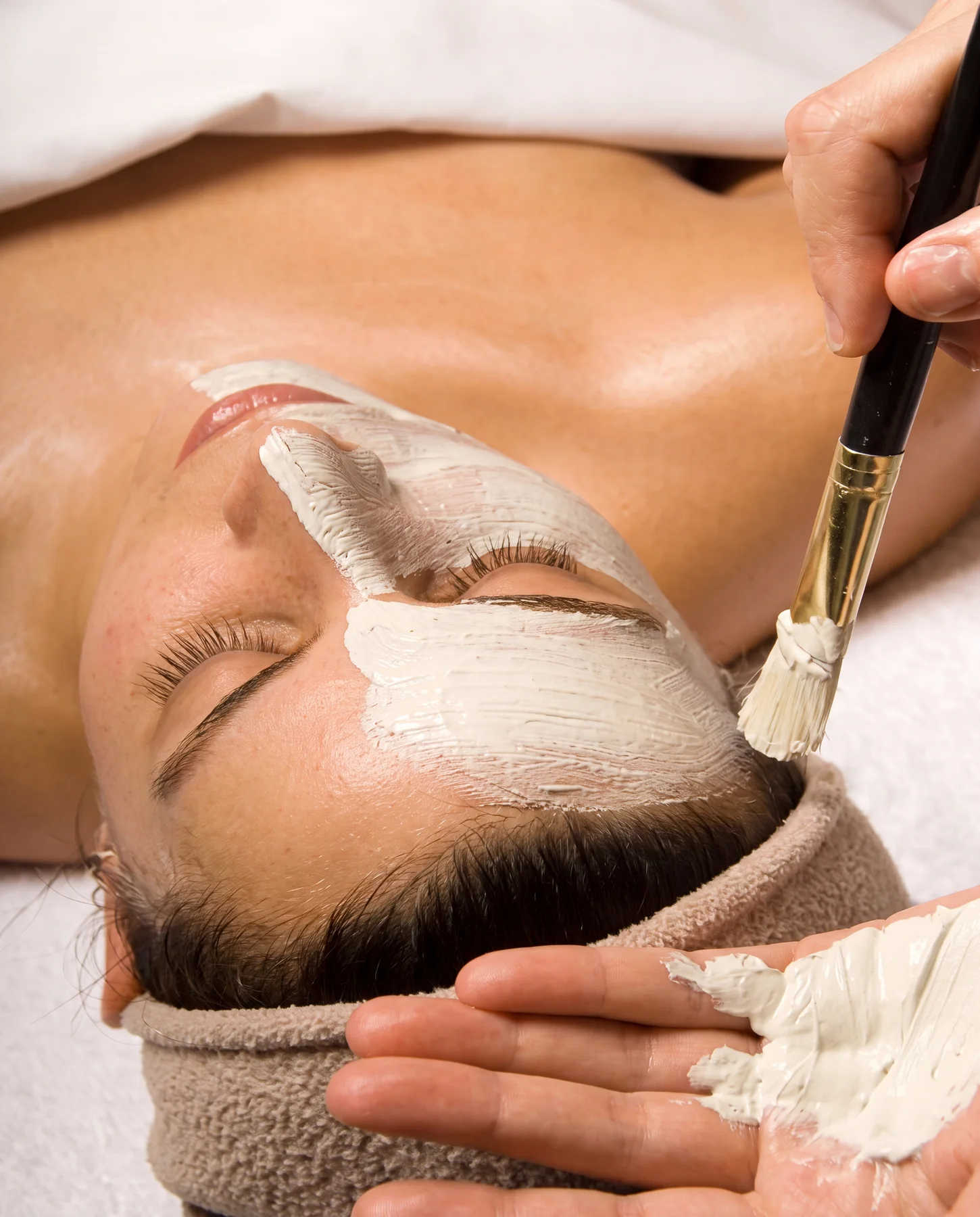 Guest resting on the facial table in a warm towel headband while her esthetician brushes on a creamy white clay mask at ESSpa Kozmetika